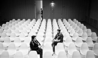 Kino Asyl 2022; ein schwarz-weiß Foto. zwei Perosnen sitzen in einem Raum voller weißer Stühle