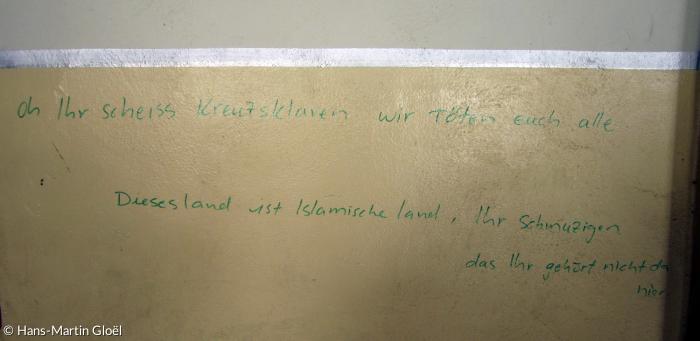 Graffito eines IS-Kämpfers mit Deutschkenntnissen in einer Seitenkapelle der Kirche von Batnaya: »Oh Ihr scheiss Kreuzsklaven wir töten euch alle. Dieses land ist Islamische land, Ihr Schmuzigen, das Ihr gehört nicht da hier«.