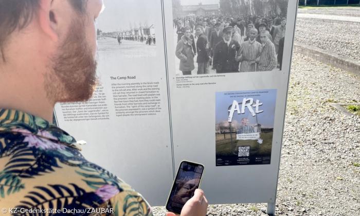 Ein Mann steht vor einer Tafel. Auf der Tafel ist ein Plakat zur Aktion ARt zu sehen. Der darauf abgebildete QR Code wird gerade von seinem Handy gescanned
