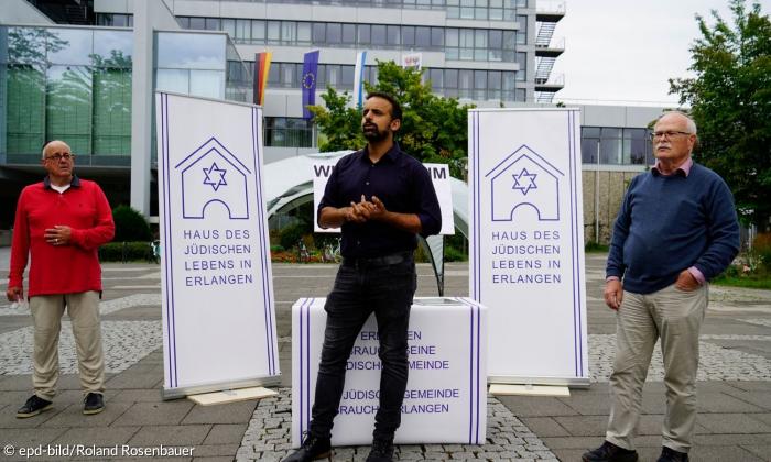 Drei männliche Mitglieder der Jüdische Kultusgemeinde Erlangen stehen auf einem steinernen Platz von Bannern mit der Aufschrift "Haus des jüdischen Lebens in Erlangen". 