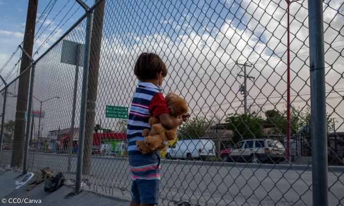 Ein Kind steht an einem mit Stacheldraht gesicherten Zaun, es hat einen Teddybär im Arm