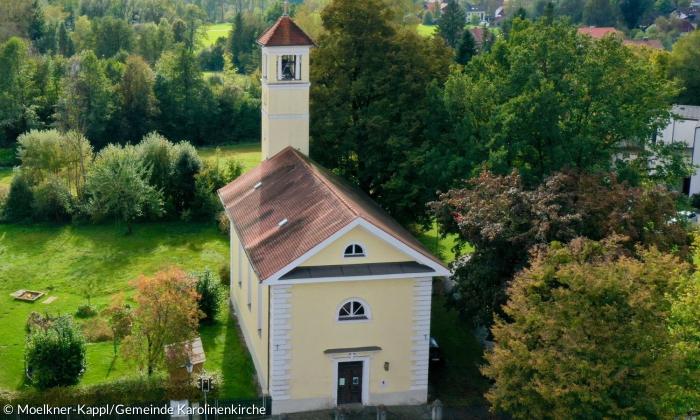 Die in einem hellen gelb gehaltene Karolinenkirche in Vogelperspektive. Die Kirche hat ein Satteldach und einen Kirchturm.