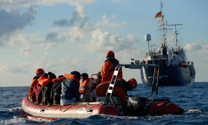 Menschen mit Rettungswesten auf einem Schlauchboot, das vor einem großen Schiff treibt.