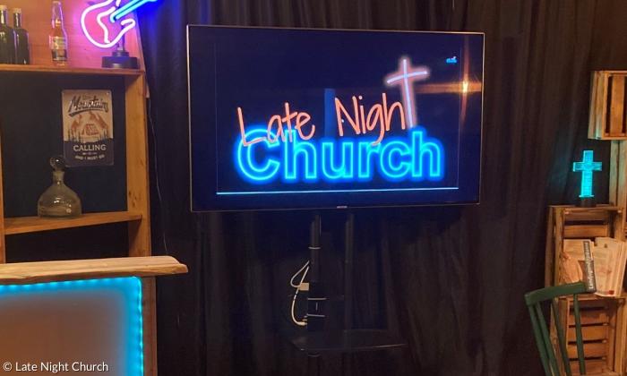 Das Studio der Late Night Church. Es stehen rustikale Regale herum, an der Wand hängt ein schwarzer Vorhang, davor ein Fernseher, auf dem das Logo der Late Night Church zu sehen ist.