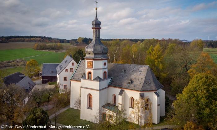 Das Gebäude der Markgrafenkirche Kirchgattendorf aus der Luft. Sie hat einen Kirchturm mit einer Uhr und besteht sonst aus einem nahezu viereckigen Hauptgebäude. Im Hintergrund sind Felder und herbstliche Bäume zu sehen.