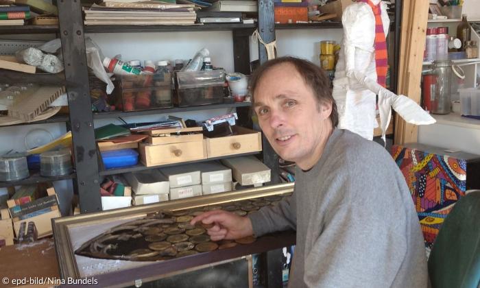 Ein Mann sitzt in dem Atelier eines Künstlers, umgeben von einer Gipsfigur und Farben.