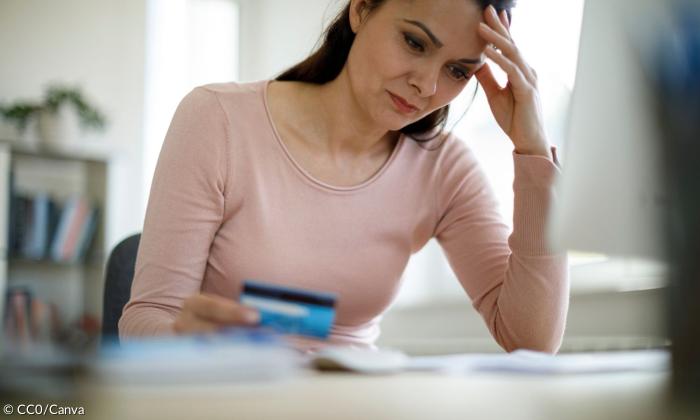 Eine Frau sitzt an einem Tisch und stützt den Kopf in die Hand, in der anderen Hand hält sie eine Kreditkarte