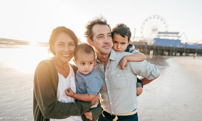 Ein Symbolbild für Urlaub. Eine Familie steht am Strand. Die Mutter hat einen Sohn im Arm, der Vater einen Huckepack. Im Hintergrund sieht man ein Pier mit einem Riesenerad.