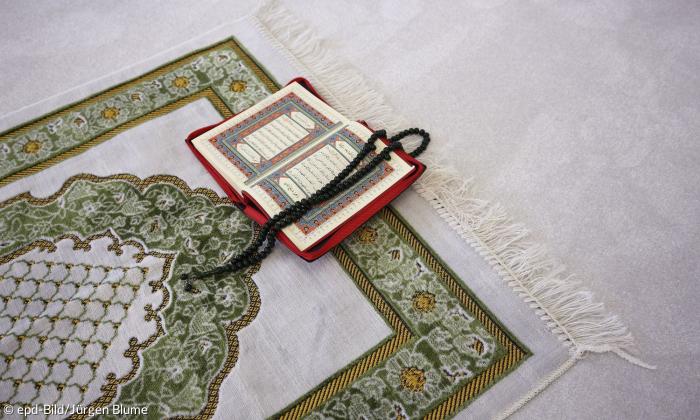 Ein aufgeschlagenes Buch liegt auf einem Teppich. Eine Gebetskette mit Perlen liegt auf dem Buch.