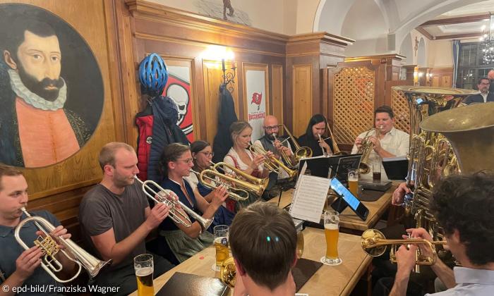 Zehn Personen sitzen in einem Wirtshaus um einen Tisch. Sie spielen Blechblasinstrumente. Trompeten und Tuba. 