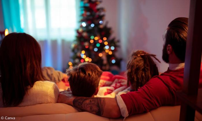Eine Familie sitzt in ihrem Wohnzimmer auf der Couch und schauen auf einen vor ihnen aufgestellten Christbaum, der bunt leuchtet. Die Eltern schließen zwei kleinere Kinder in ihre Mitte, wobei der Vater seinen Arm um sie schlingt.
