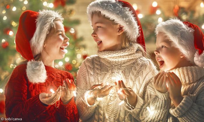 Drei Kinder haben Weihnachtsmützen auf dem Kopf und eine Lichterkette in der Hand und lachen sich an 