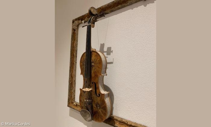 Eine Geige hängt an der Wand in einem Bilderrahmen. Es sind Blüten auf dem Geigendeckel 