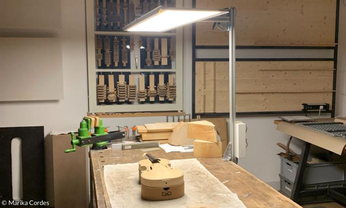 Werkbank, eine flächige Lampe beleuchtet einen rohen Geigenkörper, der auf dem Tisch liegt. An der Wand viel Holz.