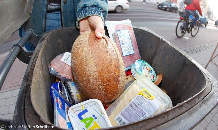 Eine Hand wirft Brot in einen Mülleimer, in dem schon Lebensmittel liegen