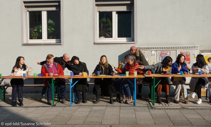 Mehrere Menschen unterschiedlicher Kulturen sitzen an einem langen Tisch, der auf der Straße steht. Sie diskutieren lebhaft miteinander und sitzen alle auf einer Seite des Tisches.