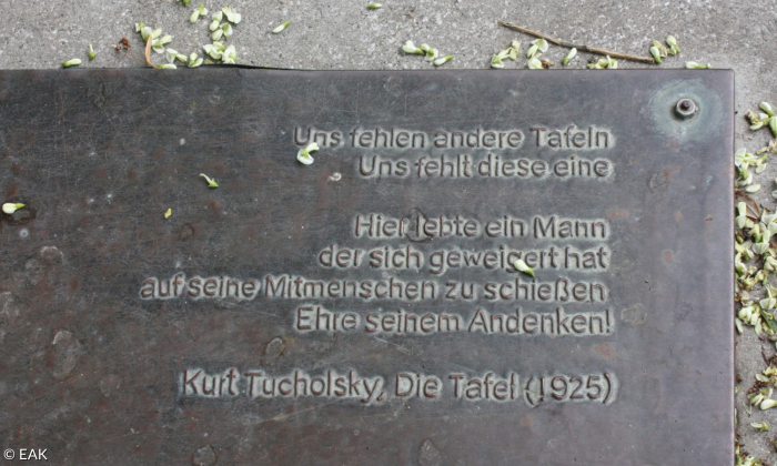 Eine Gedenktafel liegt auf dem Boden. Am oberen und rechten Bildrand liegen ein paar hellgrüne Blütenblätter herum. Auf der Tafel steht der Text "Die Tafel" von Kurt Tucholsky