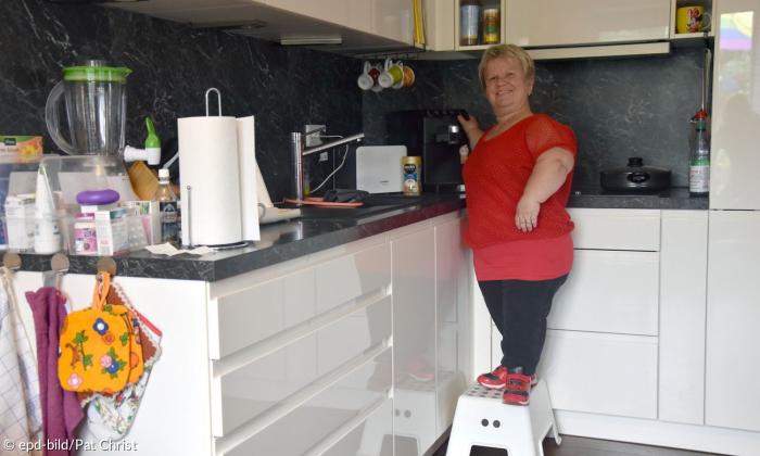 Eine kleinwüchsige Frau mittleren Alters steht in einer Küche auf einer kleinen Leiter. Sie bedient gerade die Kaffeemaschine und lacht in die Kamera. 