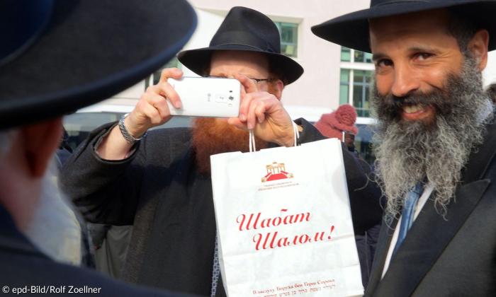 Szene zeigt einen Rabbiner mit langem Bart, der in die Kamera lächelt. Neben ihm steht ein anderer, der mit einem Handy filmt. 