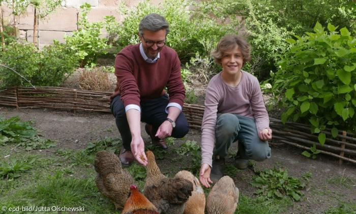 Ein Vater und sein Sohn, die bei beide im Vorgarten einer Kirche knien. Vor ihnen sind fünf Hühner, die von den beiden gefüttert werden. 