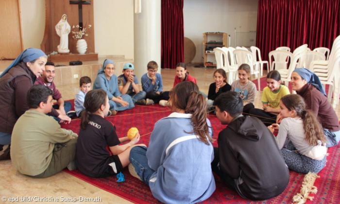 Man sieht einen Sitzkreis von Kindern auf einem roten Teppich in einem kleinen Saal. Zwischen ihnen sitzen zwei Lehrerinnen, die mit den Schülerinnen und Schülern interagieren.