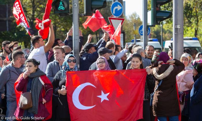 Man sieht eine Kundgebung von ungefähr hundert Menschen, die auf einer Straße stattfindet. Frauen, Kinder und Männer halten dabei Türkei-Flaggen in die Luft. Im Hintergrund sieht man große Polizeiwägen.