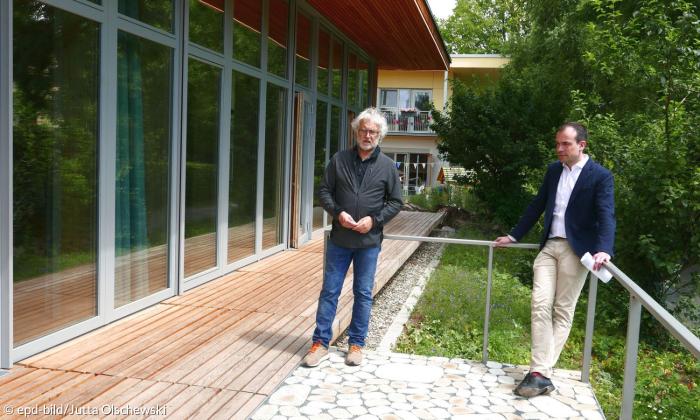 Zwei Männer stehen nebeneinander auf einer Terrasse. Links neben ihnen sieht man eine große Glasfront eines Holzhauses.