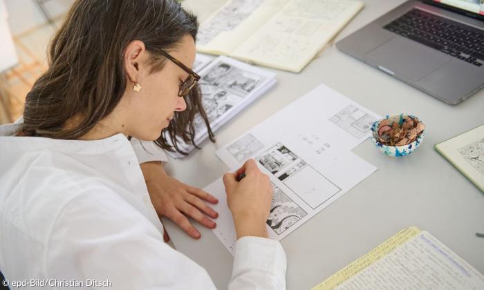 Eine Frau Mitte zwanzig sitzt gebeugt über einer Comiczeichnung. Sie hat einen Stift in der Hand und ist gerade dabei, etwas zu zeichnen. Vor und neben ihr liegen ausgebreitet beschriftete Papierblätter.