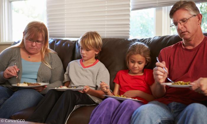 Eine Familie mit Mutter Vater und zwei kleinen Kindern auf einem Sofa und isst leblos. 