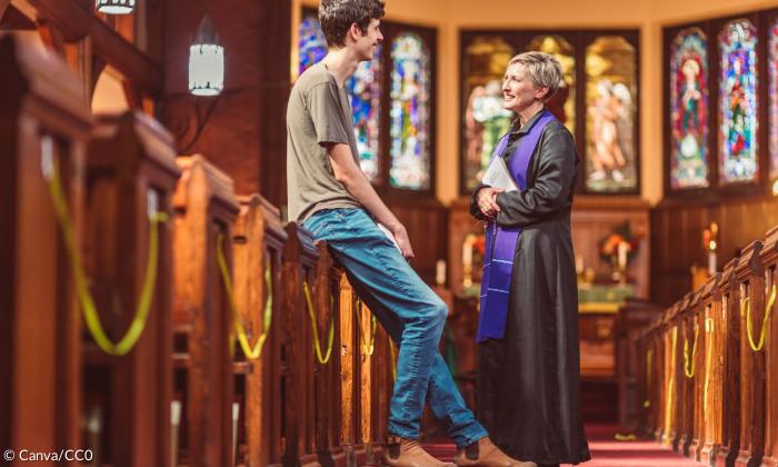 Eine Pfarrerin im Talar und lilanem Schal unterhält sich in einer Kirche mit einem jungen Mann. Sie stehen auf dem Hauptweg, der zwischen den Bänken zum Altar führt. 