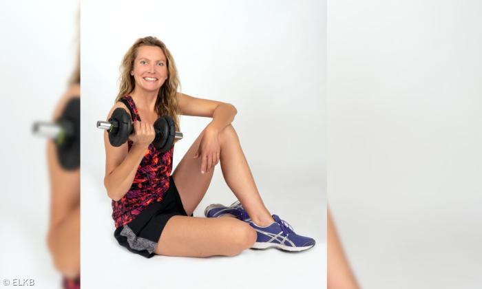 Eine Frau Anfang vierzig, die in einem sportliche Outfit lächelnd auf dem Boden sitzt. In ihrer rechten Hand trägt sie eine Hantel. 