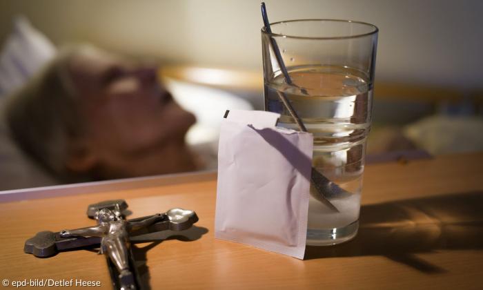 Ein Glas mit Wasser neben einer offenen Packung, daneben ein Kruzifix, dahinter eine alte Person im Bett