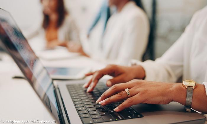 Finger tippen an einem Laptop, im Hintergrund weitere Menschen unscharf