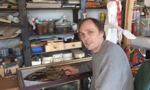 Ein Mann sitzt in dem Atelier eines Künstlers, umgeben von einer Gipsfigur und Farben.