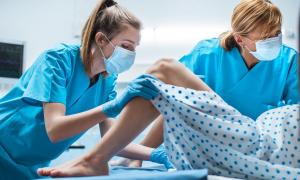 Eine junge Frau mit steriler Kleidung und einer Maske steht vor den angewinkelten Beinen einer liegenden Frau in einem Patientenkittel.