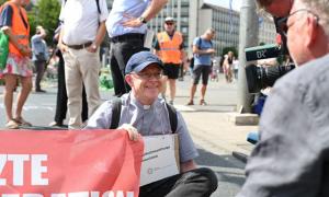 Ein Pater sitzt auf einer Straße und wird vom bayrischen Rundfunk gefilmt. Mit der rechten Hand hält er ein Banner. Hinter ihm: Menschen in Warnwesten und Passanten.