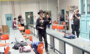 Eine Mann mit schwarzem Hut steht in einem Labor, um ihn herum Menschen mit Kameras