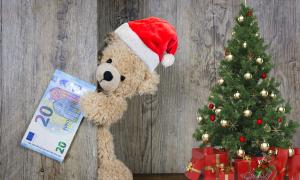 Ein Teddybär schaut aus einer Türe raus und hält einen 20 Euro Schein. Er trägt eine Weihnachtsmütze. Rechts von ihm steht ein kleiner Baum, der mit roten und goldenen Kugeln geschmückt ist. Darunter liegen Geschenke, die in rotes Geschenkpapier eingepackt sind.