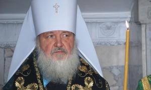 Der russisch-orthodoxe Patriarch Kyrill trägt eine weiße Haube mit einem Kreuz vorne drauf. Der Mann hat einen langen weißen Bart und eine dunkle Scherpe, die golden bestickt ist. Recht neben ihm brennt eine kerze.