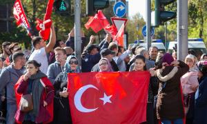 Man sieht eine Kundgebung von ungefähr hundert Menschen, die auf einer Straße stattfindet. Frauen, Kinder und Männer halten dabei Türkei-Flaggen in die Luft. Im Hintergrund sieht man große Polizeiwägen.