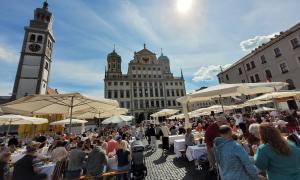 Mehrere Hundert Menschen feiern auf dem Rathausplatz in Augsburg bei strahlendem Sonnenschein an der großen Friedenstafel zusammen das Friedensfest.