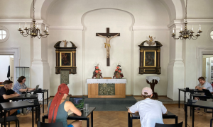 Sechs Studierende sitzen im Halbkreis and Schreibtischen vor einem Altar über de, ein kreuz hängt. Rechts und links hängt jeweils ein Kronleuchter.