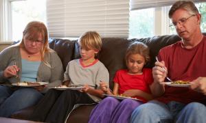 Eine Familie mit Mutter Vater und zwei kleinen Kindern auf einem Sofa und isst leblos. 